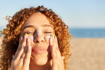skin-care-tips a smiling happy woman applying sunscreen to her face on the beach, having read our expert skin care tips for living abroad