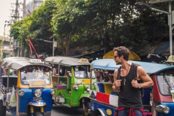 tuk-tuk-thailand a male tourist standing next to tuk-tuks in Thailand, aware of potential travel scams like overcharging tourists in popular destinations