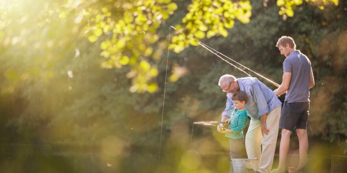 grandad-fishing-with-gandsons an eldery man sitting on a pier by a lake fishing with his two grandsons