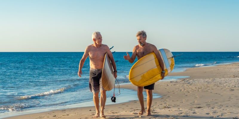 Two mature men walking with surfboards on beautiful beach enjoying paradise and retirement lifestyle. Attractive fit senior adults friends having fun surfing. In real active and healthy people. Senior Austrlaian surfers need overseas health cover