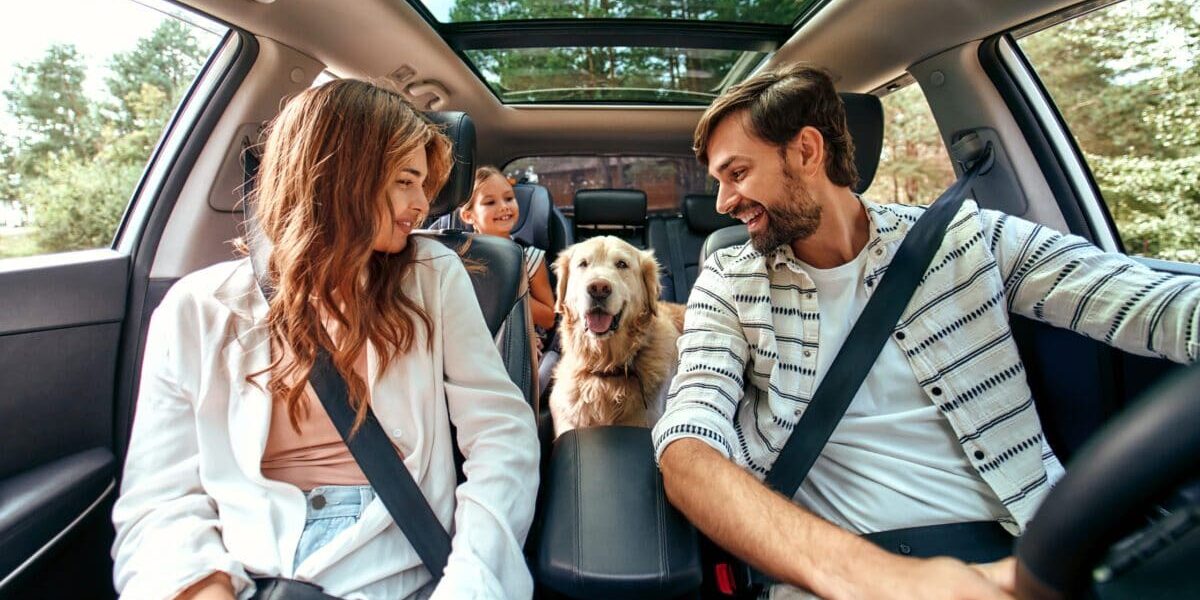 Family with dog in the car family traveling in a rental car and smiling at their dog, who they know is covered by their single-trip travel insurance plan