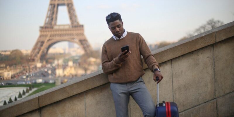 man-by-eiffel-tower a Bahamian man standing in front of the Eiffel Tower with a suitcase, looking at his phone to check the wording of his insurance for Bahamian citizens